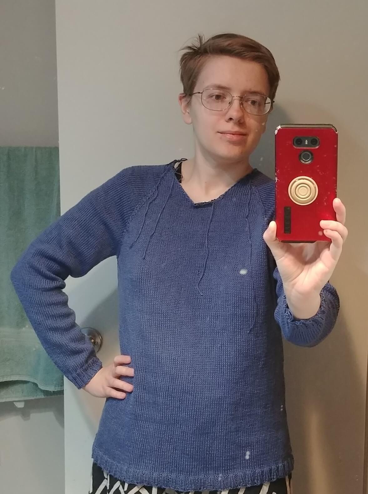 Bathroom mirror selfie of me wearing a dark blue stockinette knit sweater with ribbed cuffs and hem. The neck hole is unfinished and has bits of yarn dangling off it. 