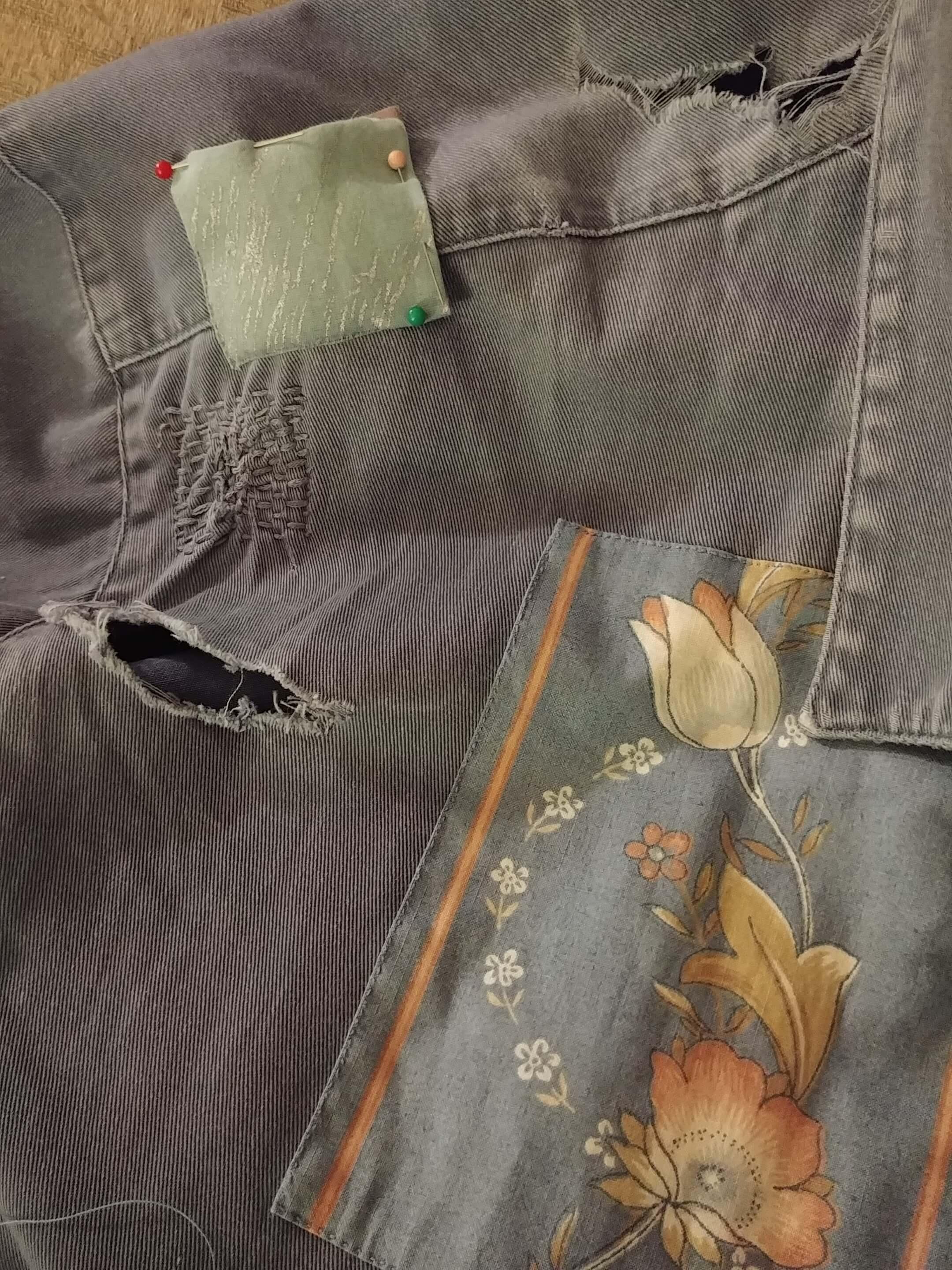 Closeup of a blue jacket with several large rips, a large floral patch, some darning, and a smaller blue and silver patch that's pinned to the jacket and partially sewn down