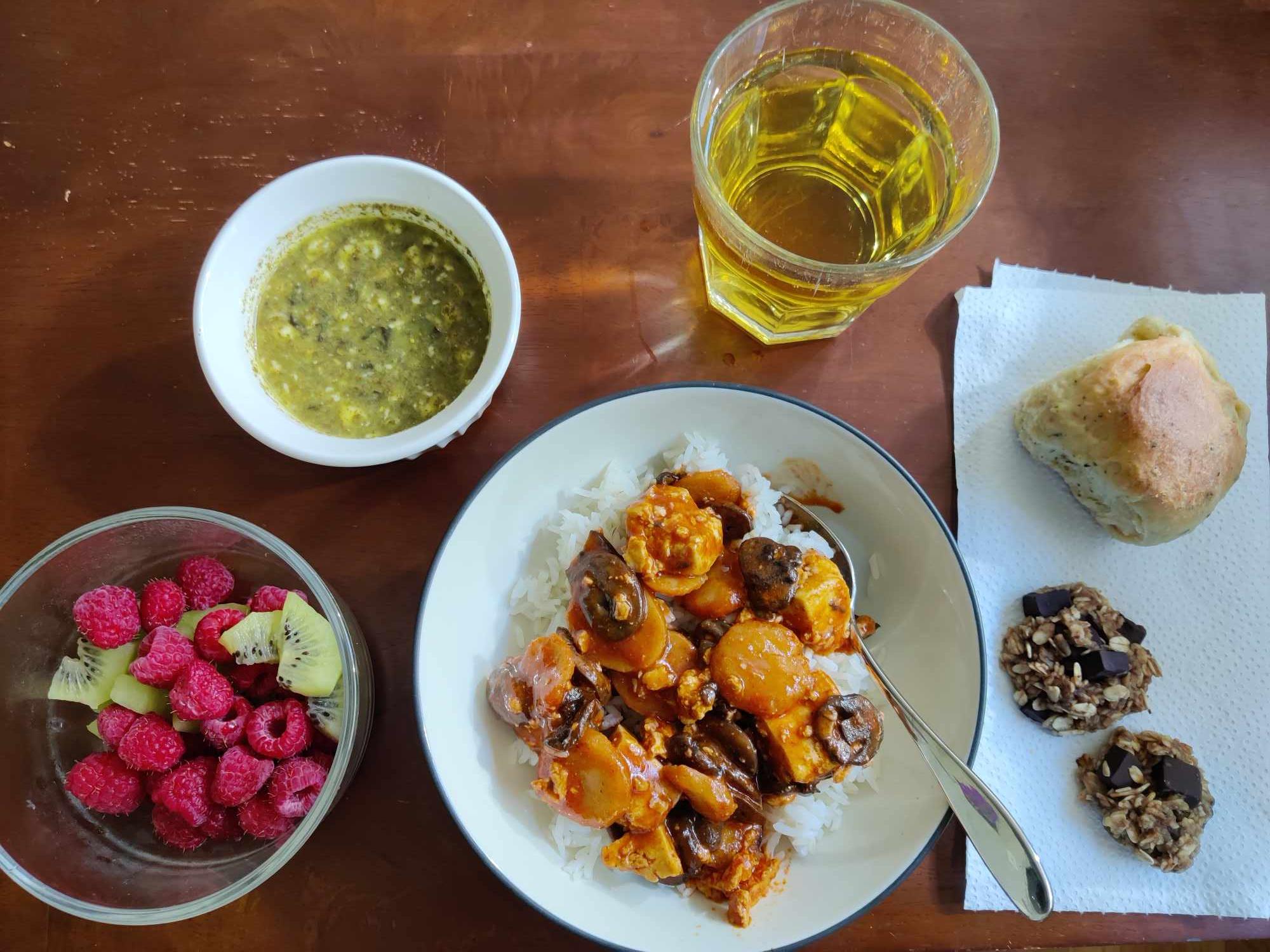 A fruit salad including raspberries and kiwis, a small bowl of pesto dip, a bowl of white rice with tofu and vegetables in an orange sauce, a glass of apple juice, a bread roll, and two small chocolate chip oat cookies