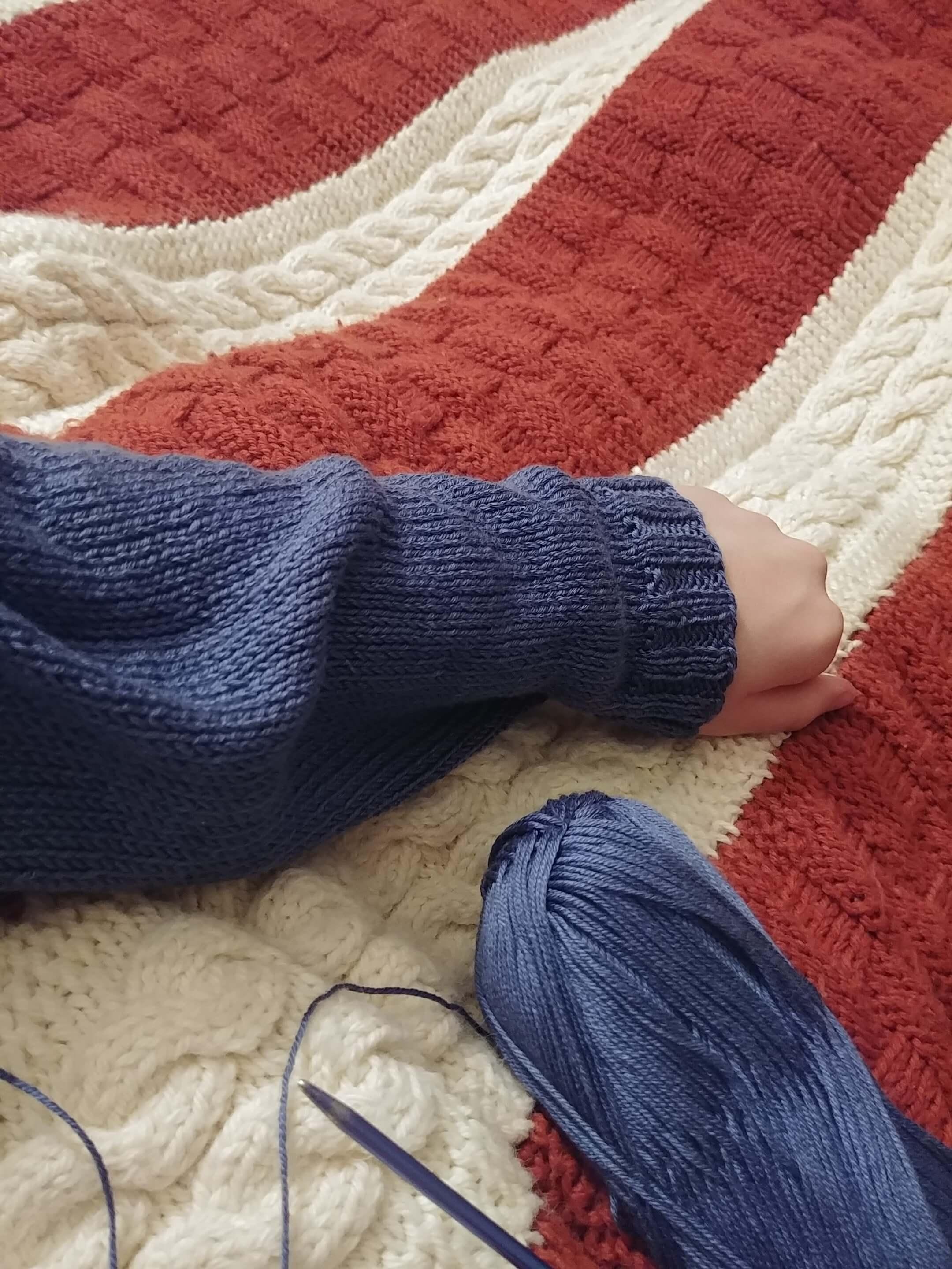 My forearm is wrapped in a blue knit sweater sleeve in progress, resting on a cable knit blanket.