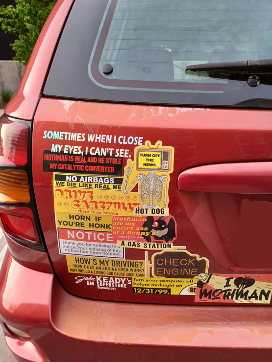 a menagerie of stickers on the back of a car, arranged such that they are overlapping
from top to bottom, left to right it states, "Sometimes When i Close my eyes, I cant see.", "Mothman is real and he stole my catalytic converter", "no airbags, we die like real me", "Drive carefully, there is no heaven", "honk if youre honk", "NOTICE, thank you for noticing this notice. Your noticing of this notice has been noticed." "How's My Driving? How does an engine even work??? Why would a loving god cause such agony??", "John Keady's GM superstore, Davenport", A  skeleton is present with a tv head that states "turn off the news", continuing below the skeleton are stickers that state "hot dog", "mothman ate my entire ass at denny's! it was a grand slam breakfast!", "A gas station", a check engine light is present, "Turn your computer off before midnight on 12/31/99, bestbuy", "I heart (in the shape of a moth" mothman"