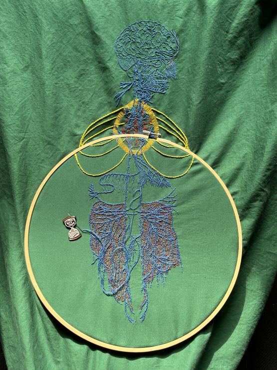 An image of an old engraving of a vagus nerve is embroidered on green cotton. The gut is in a large bamboo embroidery hoop. The embroidery of the main nerve lines is done in blue chain stitch in perlé cotton. The brain and some of the face is stitched in the same thread in backstitch. Details in the face are stitched in single strand floss. A bright red anatomical heart is stitched in red whipped back stitch. Around it a bright golden yellow halo in a vesica piscis shape is stitched in single strand thread. Three sets of ribs have are stitched in bright green/yellow stem stitch. There are some tiny details in different colours along some of the nerves. Burgundy roiling with sparse tiny black French knots are stitched in the gut area. The nerve endings in the diaphragm area are being lengthened with two strand whipped back stitch. A needle minder in the shape of an hourglass that reads, "This is taking forever," sits near the stitches holding a needle.
