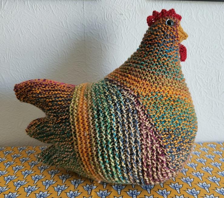 A multicoloured knitted chicken, sat on a yellow patterned tablecloth against a white wall.