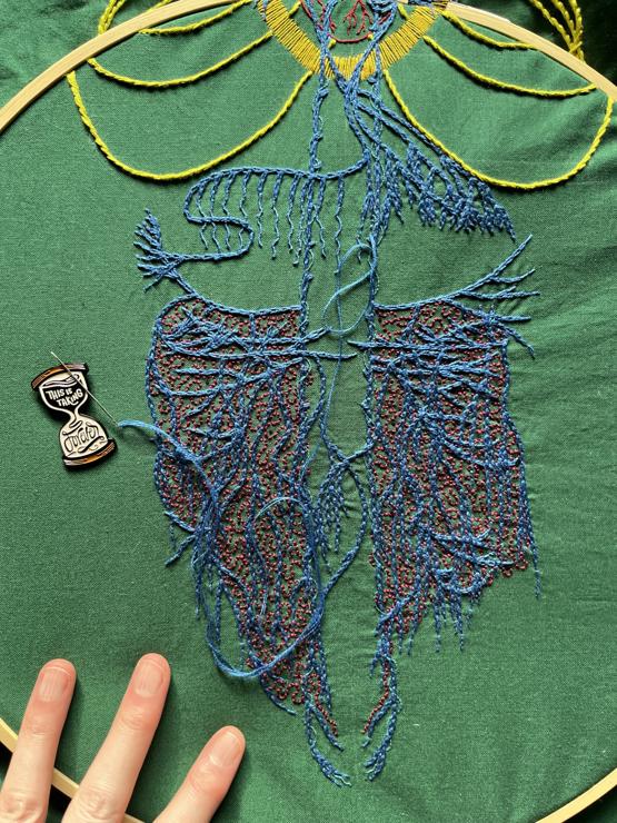 A close up of an embroidery focused on the abdominal vagus stitched in blue chain stitch in perlé thread on dark green cotton. There are many layers of crossing wavy lines. Spiralling around the very straight main vagus is single strand whipped back stitch in electric blue. Some roiling burgundy back stitch in burgundy perlé thread is between the blue. Sparse tiny black French knots are stitched in the gut area. The nerve endings in the diaphragm area are being lengthened with two strand whipped back stitch. A needle minder in the shape of an hourglass that reads, "This is taking forever," sits near the stitches holding a needle.