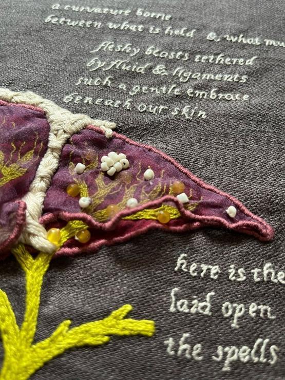 A red & purple organza liver on grey linen. The biliary system between the 2 layers is bilious green. A close up of the small lobe of the liver with small white beads on the top layer of organza represent abscesses, and round agate beads on the biliary ducts represent strictures. The text below it reads: fleshy beasts tethered. By fluid and ligaments. Such a gentle embrace. Beneath our skin.