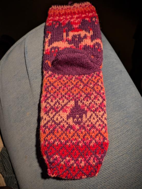 Colourwork sock. Background is pink self striping. Motif is purple. This photo shows the back of the leg and the sole. The sole has a lattice motif and a secret ghost. 