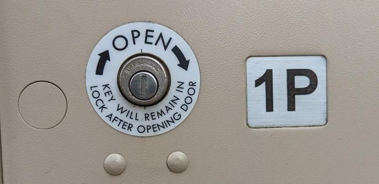 A photo of the package locker in a US shared mailbox.  A label around the keyhole reads "open" with arrows and then says "key will remain in lock after opening door" -- it's a great example of design that doesn't rely on users remembering to do the right thing (in this case, giving back the key for future use)