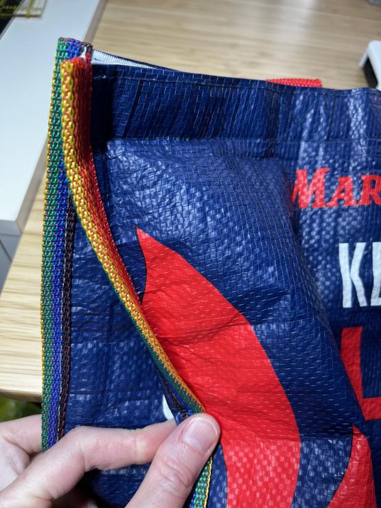 A close up view of the side of a Market Basket (local beloved grocery chain) insulated bag. The edge binding has been replaced with a strip of rainbow nylon webbing. A Caucasian hand is holding the edge so that both sides are visible. 