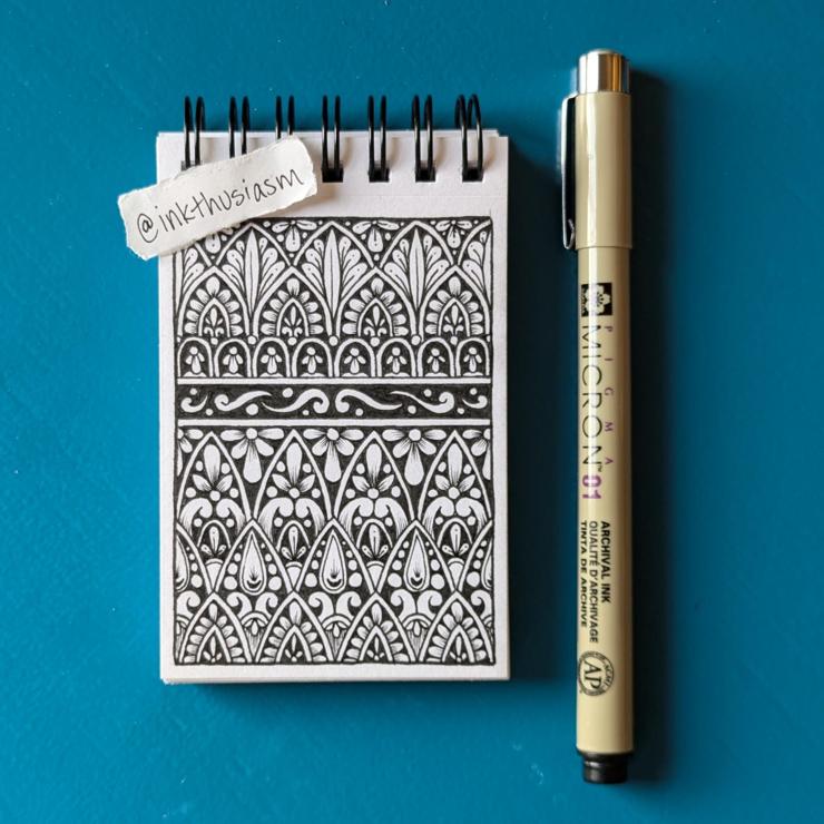 Mandala patterns are drawn in black ink in a spiral-bound sketchbook that is about the size of a business card. The Micron pen I used is next to the sketchbook, and my little handwritten "inkthusiasm" tag is placed up near the wire spirals.