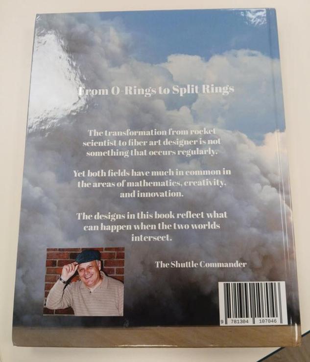 Back cover of the tatting book. It has photos of clouds and sky in the background, a small insert photo of the author who appears to be a jaunty middle aged white guy with a scally cap he's tipping. 

Text says:
The transformation from rocket scientist to fiber art designer is not something that occurs regularly.

Yet both fields have much in common in the areas of mathematics, creativity, and innovation.

The designs in this book reflect what can happen when the two worlds intersect. 

The Shuttle Commander.