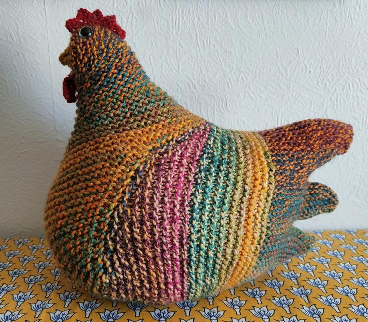 A multicoloured knitted chicken, sat on a yellow patterned tablecloth against a white wall.