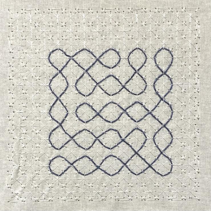 A plain beige cotton fabric contains a grid of machine-made stitches in white thread in a star-burst-like pattern. This stitched grid supports a hand-woven kolam in a navy blue single-stranded pearl cotton thread. 