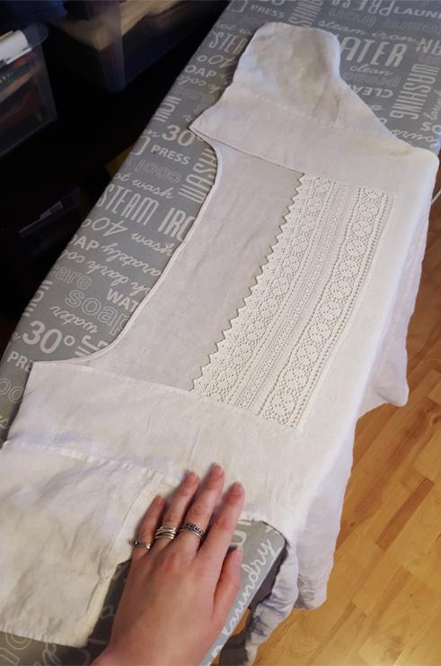 A photo of just the top part of the shift laid out on an ironing board. The front neckline has the wide and tall lace insert with a scalloped edge, the back is a curving narrow rolled hem. He neckline opening is still rather large, but not ridiculous. The sleeves of the shift are very short and kind of funny looking. Sini's hand in the frame.