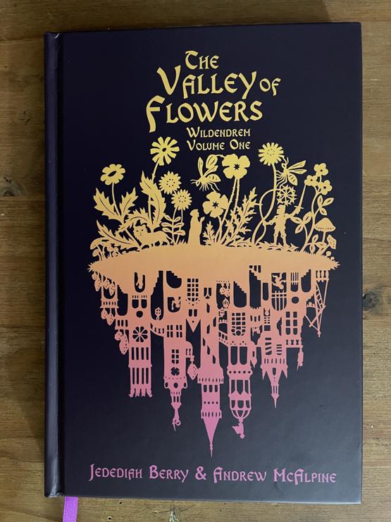 The cover of The Valley of Flowers: a gradient silhouette from yellow to pink of a group of wildflowers. Beneath them (in place of roots) are the towers of s city.