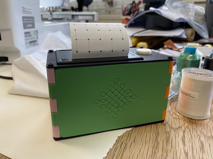 A rectangular receipt printer with a bright leafy green front panel and two side panels in pink and orange. The green front panel has a kolam engraved into it. A printed paper of a kolam line structure is emerging out of the printer from the top. The printer is sitting on a wooden table with a lot of cluttered sewing and craft materials around it. 