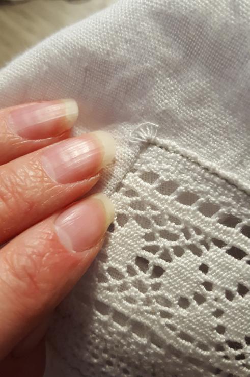 Very very close up of the reinforcing stitches on the very corners of the lace insert. They look like a miniature scallop or sea shell, made with buttonhole stitches. Sini's fingers are in the frame and the linen is such a stunning bleached white that her fingernails look quite yellow.