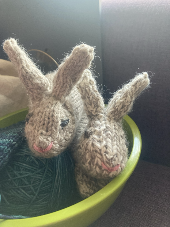 Two handknitted bunnies in natural coloured wool that approximately wild rabbits’ speckled colouring. They are facing the camera, perched on the edge of a green bowl with a teal knitted sock in progress in it.