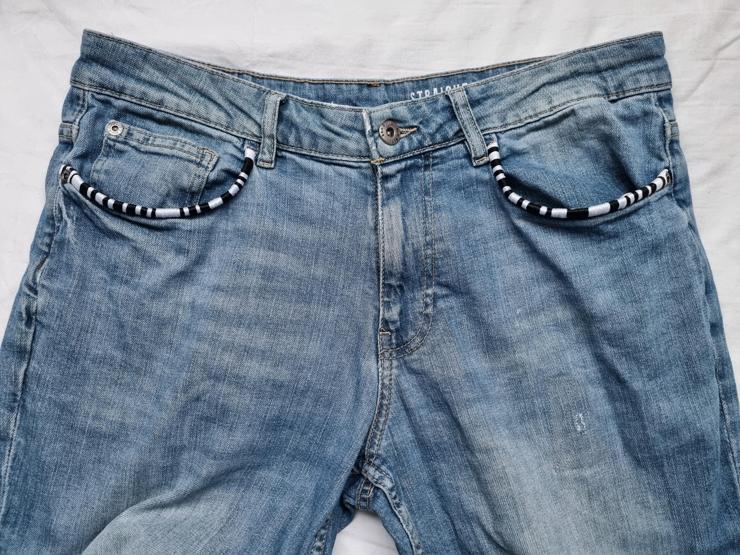 A photograph of the front of a rather worn pair of jeans. Both front pocket openings have been bound in black and white thread. The pattern is different on each pocket. 